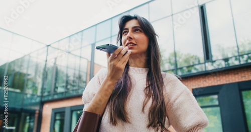 Beautiful Caucasian girl recording voice message on smartphone. Talking to microphone while holding mobile device horizontally with one hand. Thinking what to say to friend or relative. photo