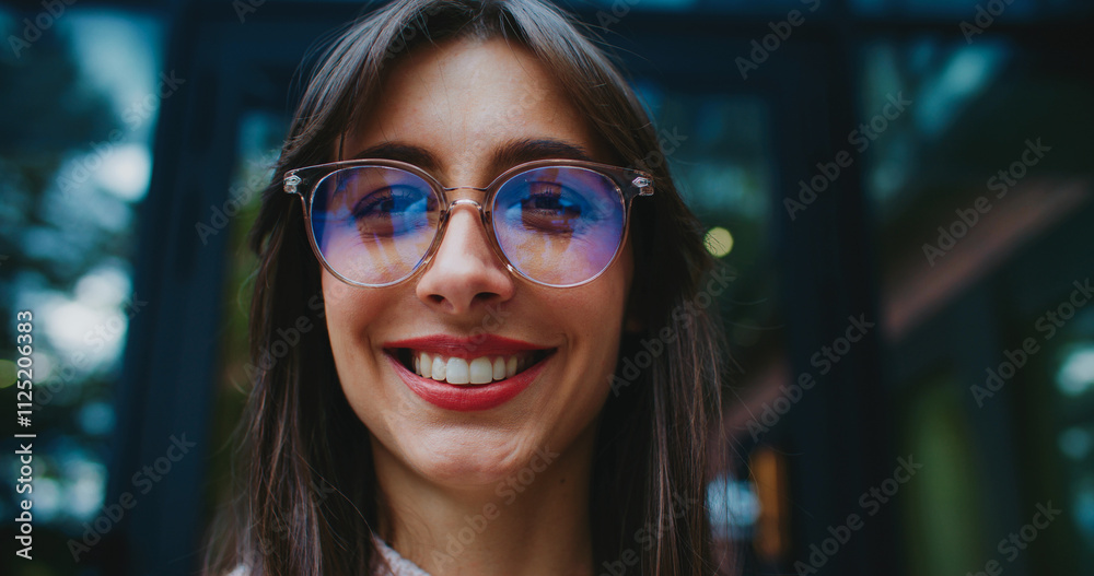 Very close facial view of Caucasian woman with round glasses looking at something on side. Glancing directly at camera. Showing warm smile on face. Trees visible in tall windows in background.