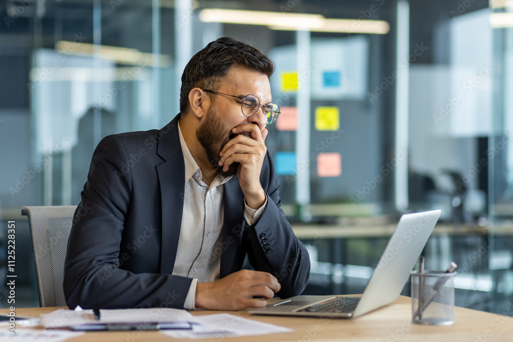 A professional man yawning as he works on his laptop, showcasing fatigue in a contemporary office setting. The image highlights workplace stress and the need for breaks during busy workdays. A professional man yawning as he works on his laptop, showcasing fatigue in a contemporary office setting. The image highlights workplace stress and the need for breaks during busy workdays.