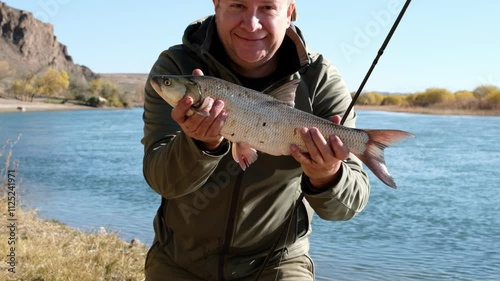 A fisherman holds a fish in his hands and rejoices at his catch. An adult fisherman stands with an asp and shows it to the camera, the fish moves in the frame, the man kisses the fish. photo