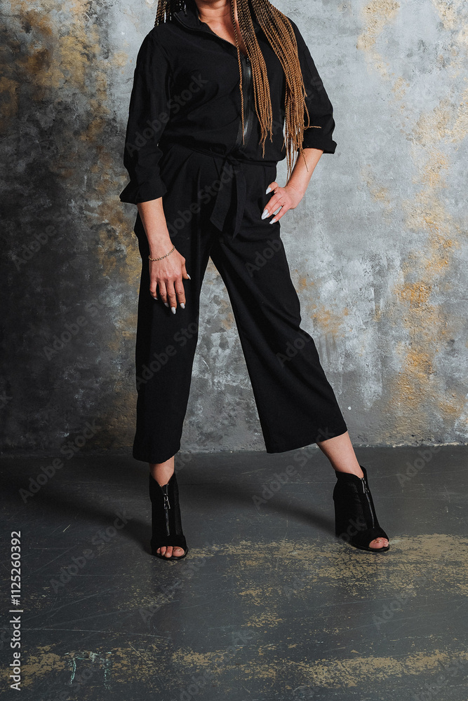 A woman wearing a sleek black jumpsuit is standing confidently in front of a clean wall, showcasing her fashion sense and posing for a striking photo shoot in this stylish outfit A woman wearing a sleek black jumpsuit is standing confidently in front of a clean wall, showcasing her fashion sense and posing for a striking photo shoot in this stylish outfit