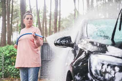 Mid age woman with bun rinsing her car with a blue hose used to remove soap and dirt on her home driveway