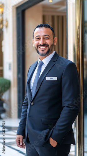 A cheerful male concierge stands confidently in a smart suit, smiling warmly at the camera, embodying hospitality in an elegant hotel setting