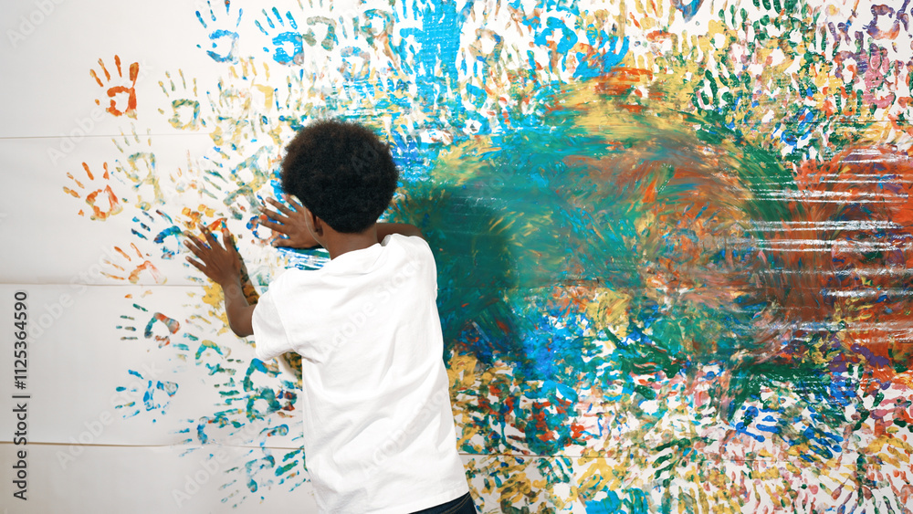 Back view of happy african boy painted the stained wall with colorful hand while wearing casual white shirt in art lesson.Smart student use hand print to make creative artwork. Education. Edification. Back view of happy african boy painted the stained wall with colorful hand while wearing casual white shirt in art lesson.Smart student use hand print to make creative artwork. Education. Edification.