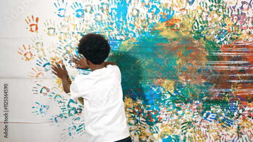 Back view of happy african boy painted the stained wall with colorful hand while wearing casual white shirt in art lesson.Smart student use hand print to make creative artwork. Education. Edification.