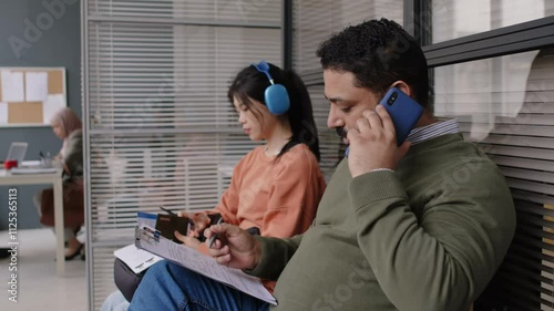 Side shot of male Biracial visa applicant in cardigan consulting how to fill out application form talking on phone while sitting in waiting room before interview in consulate
