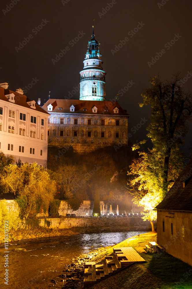 A stunning night view of the illuminated tower of Cesky Krumlov Castle towering above the Vltava River, creating a magical atmosphere with the reflection in the water