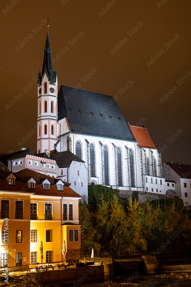 A night view of the Church of St. Vitus in Cesky Krumlov, beautifully illuminated by lights, standing tall against the dark sky, highlighting its architectural details