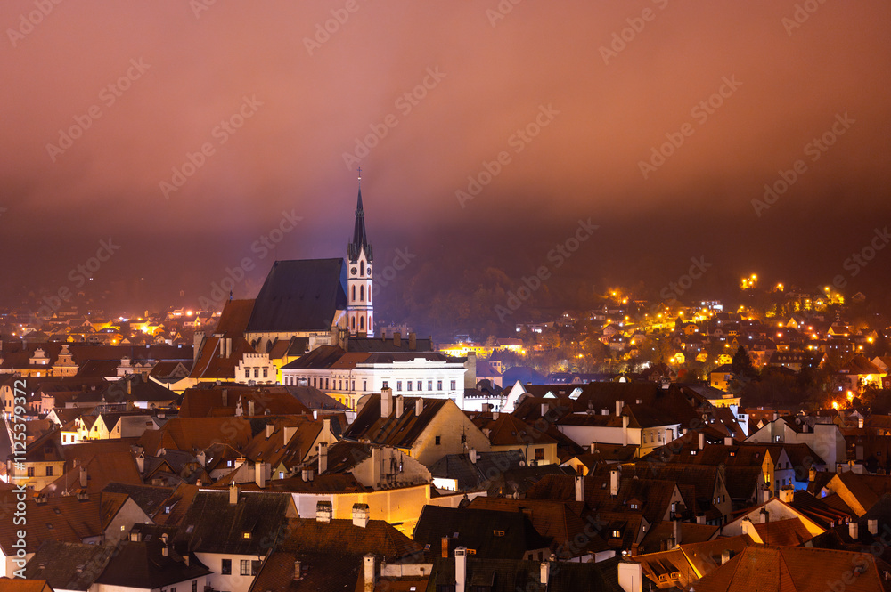 A panoramic night view of Cesky Krumlov, showcasing the rooftops, illuminated buildings, and the Church of St. Vitus beautifully lit up against the dark sky