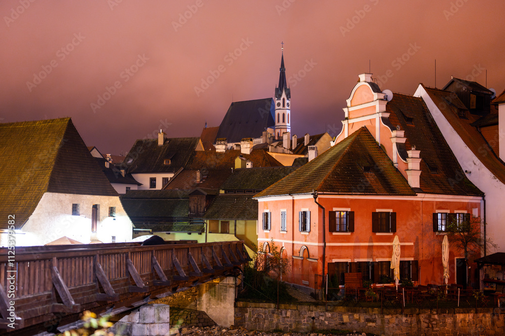 A panoramic night view of Cesky Krumlov, showcasing the rooftops, illuminated buildings, and the Church of St. Vitus beautifully lit up against the dark sky