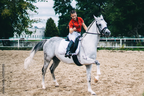 Side view of carefree Caucasian female jockey during horseback training with horsewhip in paddock spending weekend for practicing hobby and make dressage of champion stallion, equestrian sport photo