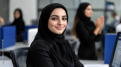 A photo of an Emirati woman sitting in front of her computer, smiling at the camera, with one other woman behind her clapping and standing beside their computers. 
