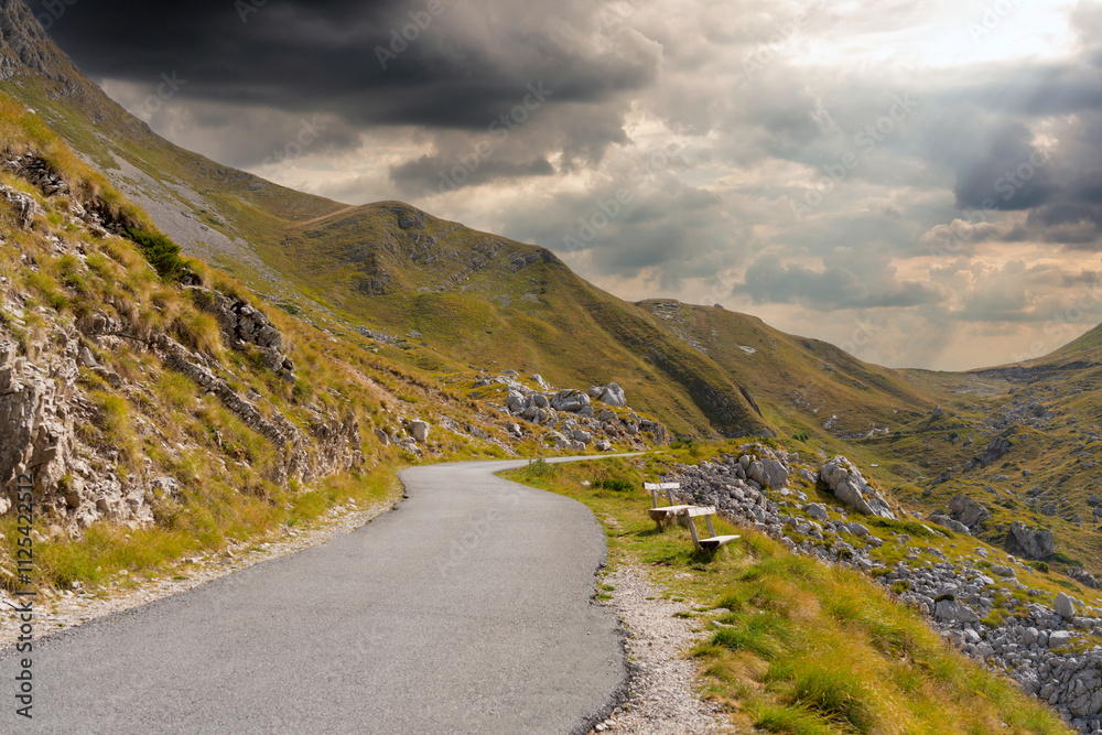 Winding mountain road through green slopes under a dramatic cloudy sky. Winding mountain road through green slopes under a dramatic cloudy sky.