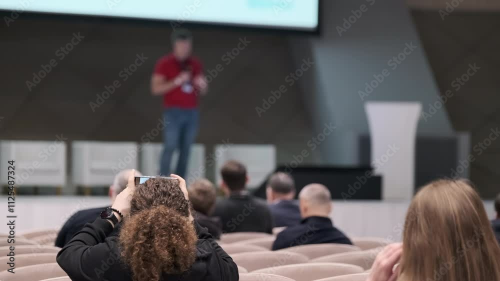 Attendees photograph presenter during technology conference. Focus on audience using phone to capture moment.
