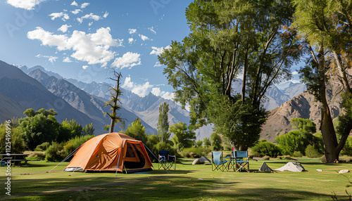 big brown dome camping tent in a green grass with trees and mountains. Taken in an afternoon with a clear blue sky with white clouds. There're chairs, table and camping equipments nearby