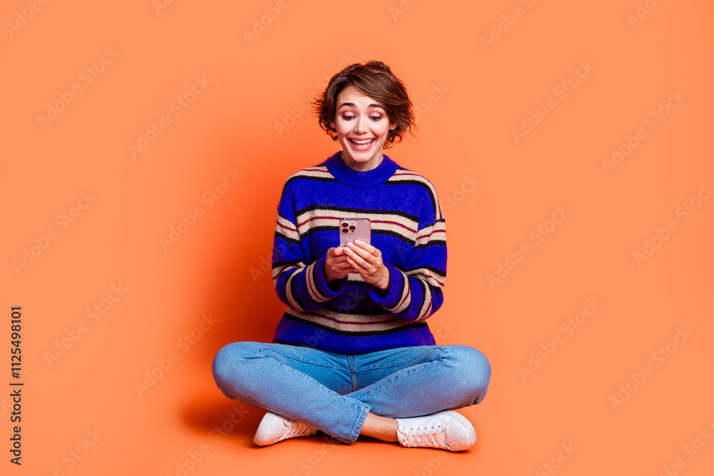 Photo of charming young girl brown hair wear striped sweater and jeans blogger just chatting in smartphone isolated on orange color background Photo of charming young girl brown hair wear striped sweater and jeans blogger just chatting in smartphone isolated on orange color background