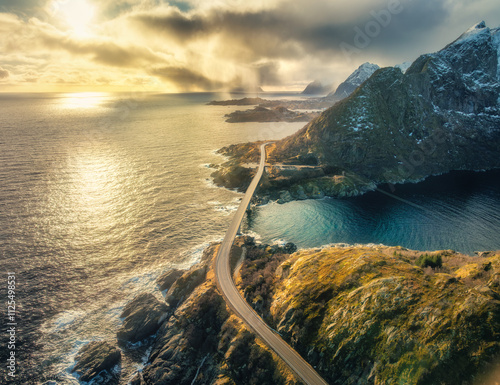 Aerial view of Djupfjord bridge crossing the fjord with snowy mountains, colorful sky in golden hour in winter. Moskenes, Lofoten islands, Norway. Top view of road, snowy rocks, blue sea at sunset photo