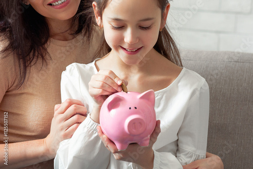 Canvas Print Happy mother and her cute teen daughter putting coin into piggybank, saving money at home, copy space