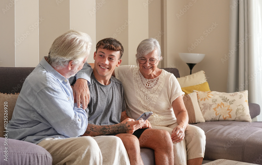 Fototapeta premium Happy multigenerational family of white-haired grandparents and young grandson sitting together on sofa at home. The old and new generation share pleasant moments in harmony