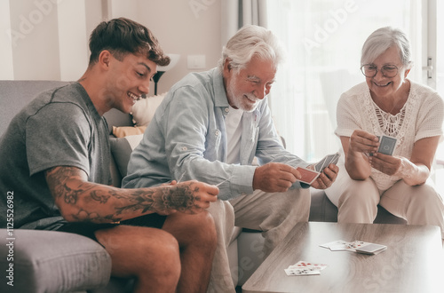 Happy multigenerational family of white-haired grandparents and young grandson play cards sitting on sofa at home. Three generations of caucasian family photo