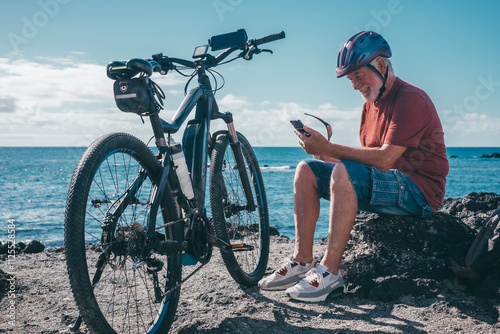 Smiling active senior cyclist man wearing helmet stops riding his bicycle to use mobile phone sitting on the beach. Authentic retirement lifestyle concept. Horizon over sea photo