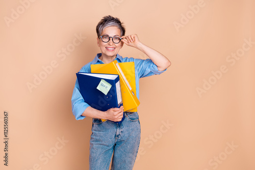 Charming senior businesswoman with trendy short grey hair holding folders standing against beige background