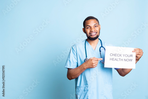 Portrait of african american guy presenting a message on a paper, urging patients to do a medical examination, blue background. Young adult specialist does a medical advertisement. photo