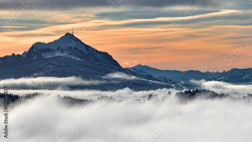 Drohnenaufnahme im Winter mit Nebel im Tal und Sonne im Gebirge im Sonnenuntergang
