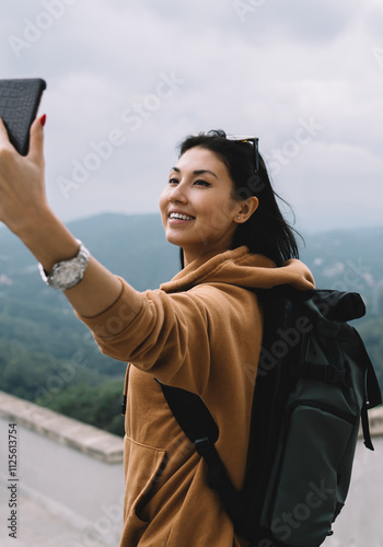 Smiling asian female tourist in casual wear and backpack making picture on mobile phone camera during vacations, cheerful woman posing for selfie exploring destinations in journey chare content online photo