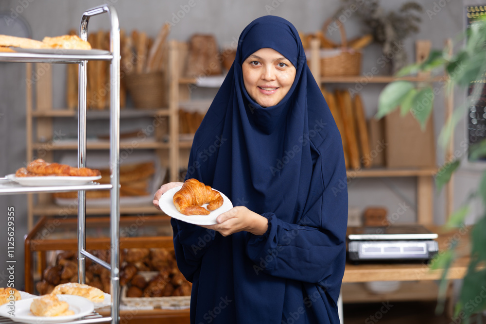 Smiling asian muslim woman holding a plate of croissant in her hands in a bakery. Woman in a hijab chooses a croissant Smiling asian muslim woman holding a plate of croissant in her hands in a bakery. Woman in a hijab chooses a croissant
