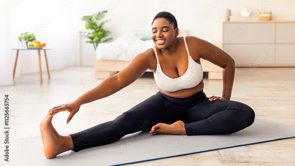 Fototapeta premium Curvy black lady stretching her legs, exercising on yoga mat, leading active lifestyle, trying to lose weight at home. Plus size young African American woman working out during her slimming program