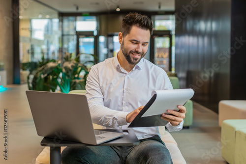 Caucasian young businessman using laptop and working with documents, analyzing information, reading papers prepare financial report. Male entrepreneur in formal analyzing graph in modern office lobby.