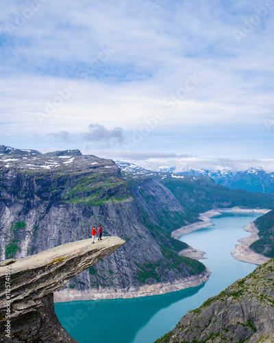 Two hikers stand on the edge of Trolltunga, Norway, a dramatic cliff overlooking a winding fjord in Norway. The sky is clear, the water is turquoise, and the landscape is breathtaking during summer