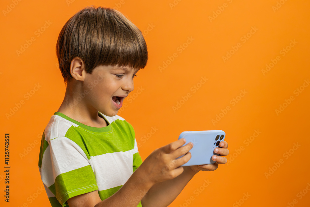 Happy excited young preteen child boy kid player celebrate victory enthusiastically playing racing drive simulator, shooter video games on smartphone. Little children isolated on orange background Happy excited young preteen child boy kid player celebrate victory enthusiastically playing racing drive simulator, shooter video games on smartphone. Little children isolated on orange background