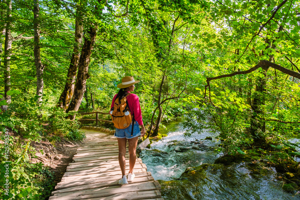 A traveler meanders along a wooden path surrounded by vibrant greenery and cascading waters, immersed in the enchanting beauty of Plitvice Lakes National Park in Croatia during summer A traveler meanders along a wooden path surrounded by vibrant greenery and cascading waters, immersed in the enchanting beauty of Plitvice Lakes National Park in Croatia during summer