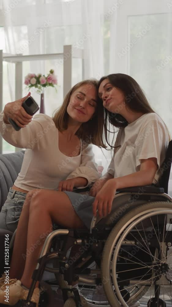 Cheerful sisters take selfies on smartphone making funny facial expressions. Woman with disability sits in wheelchair enjoying time at home Cheerful sisters take selfies on smartphone making funny facial expressions. Woman with disability sits in wheelchair enjoying time at home