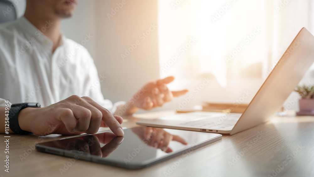 Fototapeta premium A man is using a laptop and tablet on a desk. The laptop is open and the tablet is on the table. The man is focused on the screen, possibly working or browsing the internet
