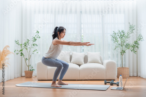 Young Asian beautiful woman doing aerobic exercise at home. Attractive girl doing  workout by squatting, follow instructions video from online trainer for health in house photo