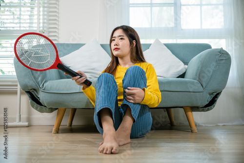 Asian female sitting floor using mosquito device swatter or electric net racket, Young woman killing mosquitoes hand holding fly swatter like weapon in living room at home, technology insect killer photo