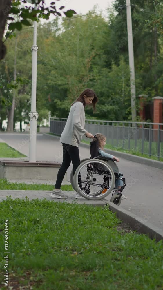 Mother carefully lowers wheelchair with daughter on road on spring day. Woman and girl with chronic health condition head home after walking in park Mother carefully lowers wheelchair with daughter on road on spring day. Woman and girl with chronic health condition head home after walking in park