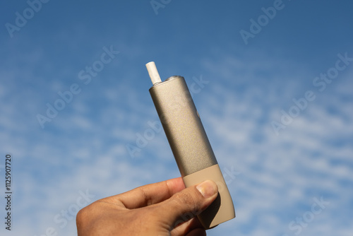 A hand holding a sleek modern heat-not-burn tobacco device against a bright blue sky with soft white clouds. Perfect for concepts of innovation, technology, smoking alternatives, and modern lifestyle.