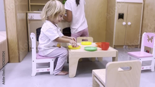 Children play in children's kitchen. Little girl in white clothe sit at small table and have fun, pretend to eat and drink. Children's games and activities at home.