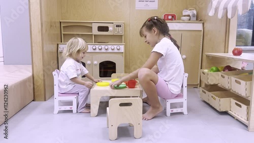 Children play in children's kitchen. Two girls in white clothes sit at small table and have fun. Children's games and activities at home.