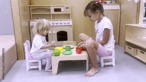 Children play in children's kitchen. Two girls in white clothes sit at small table and have fun, pretend to eat and drink. Children's games and activities at home.