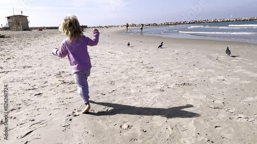 Little blonde girl runs barefoot along deserted seashore. View from back. Beach in autumn. Sea waves slowly and calmly splash.