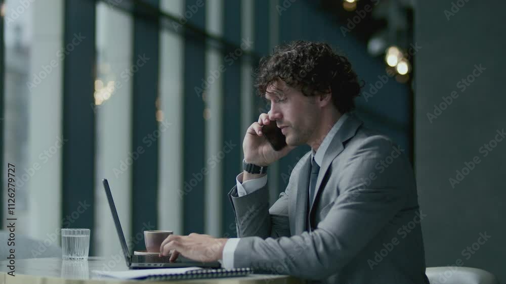 Businessman in elegant grey suit sitting at table in modern restaurant, speaking on cell phone, then working on laptop. Side view Businessman in elegant grey suit sitting at table in modern restaurant, speaking on cell phone, then working on laptop. Side view