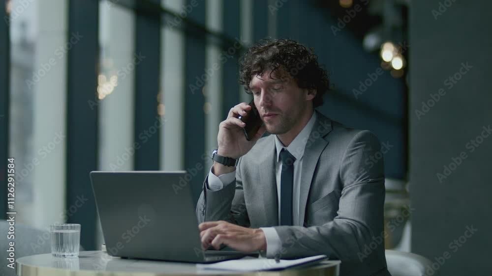 Business professional in formal grey suit sitting at table in restaurant, talking on cell phone and using laptop Business professional in formal grey suit sitting at table in restaurant, talking on cell phone and using laptop