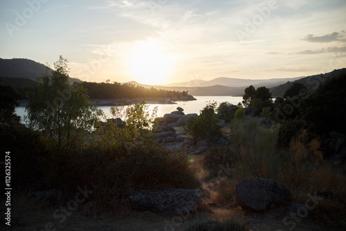 Sunset over a serene lake nestled in a valley.  Rocky shoreline, trees, and hills frame the tranquil scene. photo