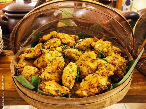 Traditional buffet with boiled bananas and fried fritters on banana leaves, covered with bamboo and cloth. Clay pot in the background adds rustic charm, highlighting Indonesian culinary heritage.