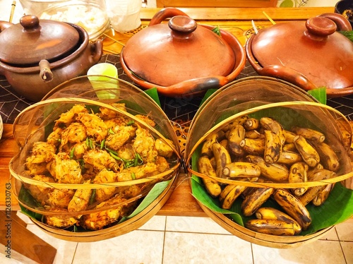 Traditional buffet with boiled bananas and fried fritters on banana leaves, covered with bamboo and cloth. Clay pot in the background adds rustic charm, highlighting Indonesian culinary heritage.